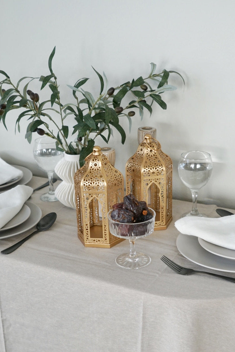 Dining table setting with gold lanterns, greenery, and white tablecloth.